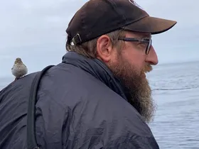 Seabird trip leader Mark Kudrav at sea, with a small landbird perched on his shoulder